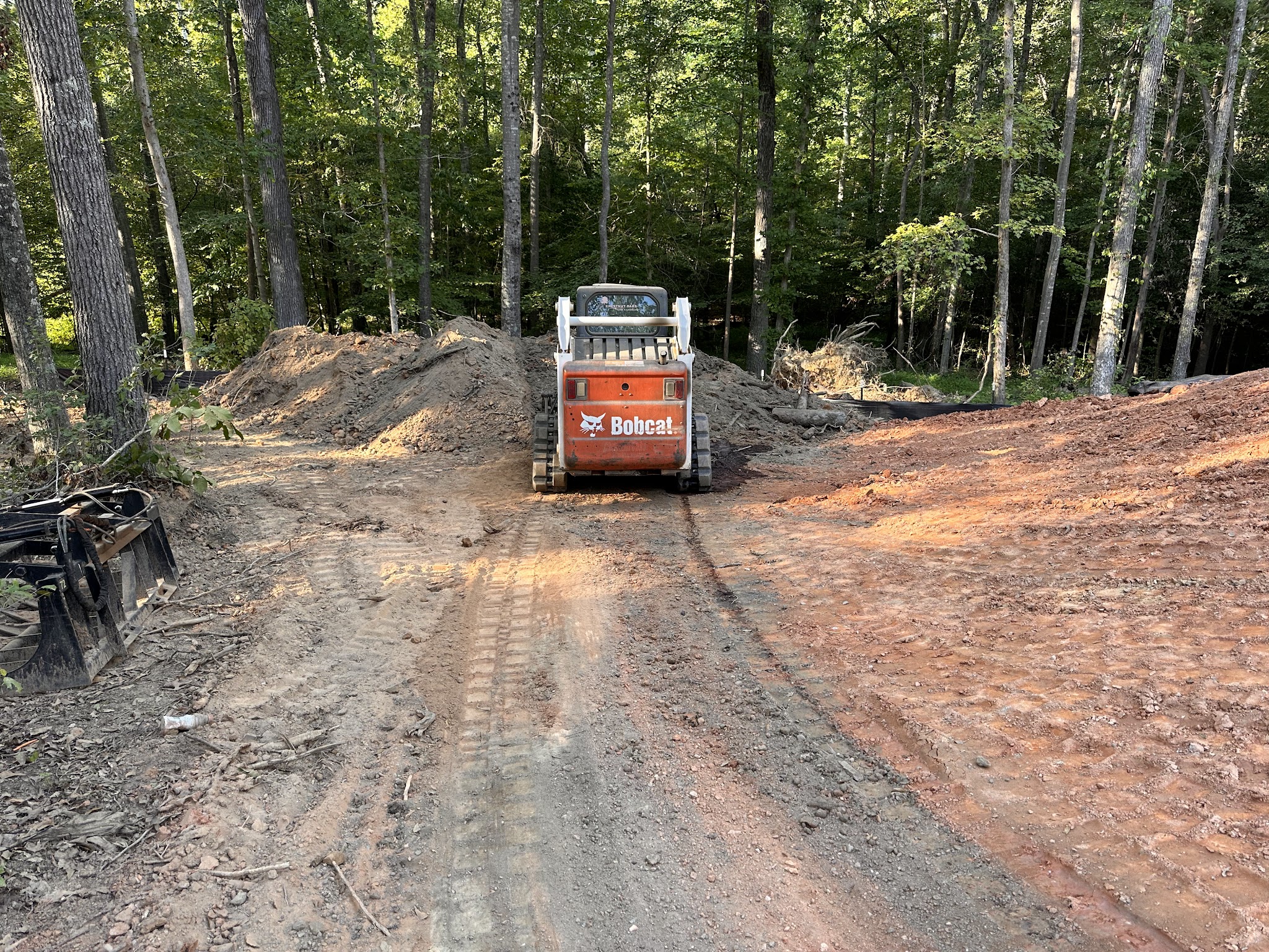 A Bobcat skid steer working on a cleared, red clay site at the edge of the woods